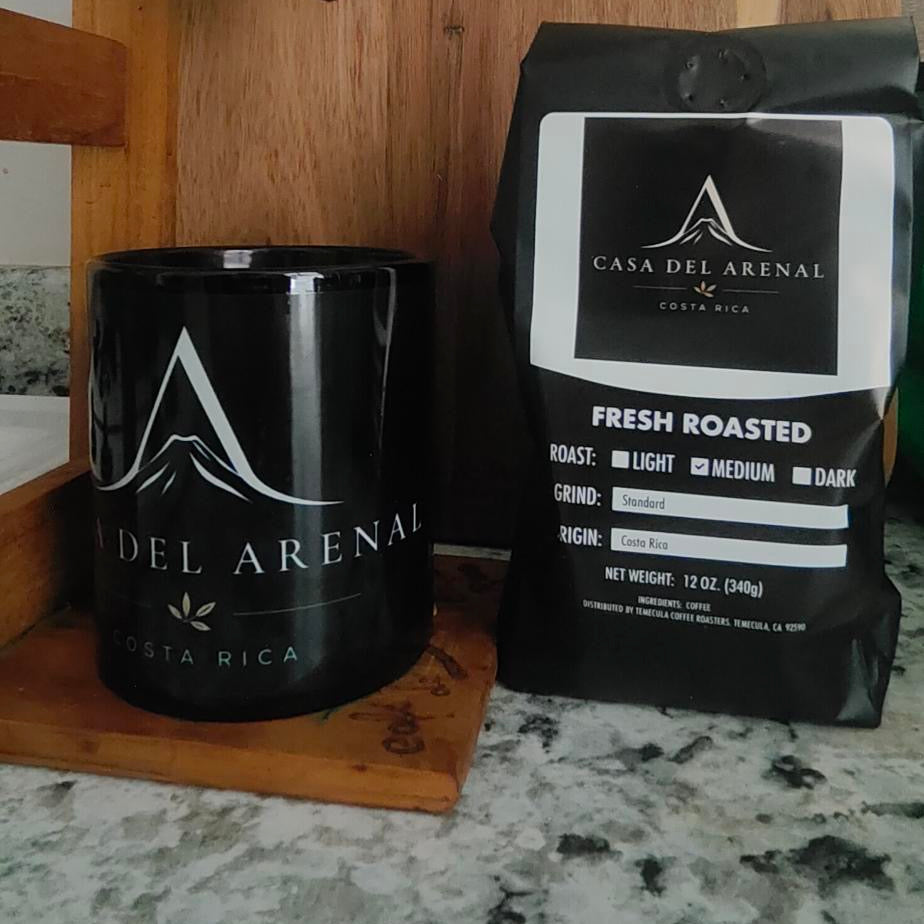 A black ceramic mug and a black bag of Casa Del Arena Fresh Roasted coffee sit on a wooden stand and a speckled gray countertop.